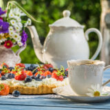 A white porcelain teapot and cup-and-saucer set arranged on a sunny table alongside a fruit tart of red and blue berries