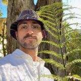 A person in a white shirt and black bucket hat poses for a photo next to a green fern frond