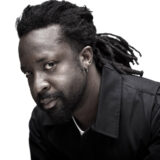 A person with long dark hair, a beard, and a black collared shirt poses for a photo against a white backdrop