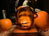 Realistic carved pumpkin face where the mouth of the pumpkin is lit up with a round light bulb. There are brown sticks bent at the sides with thin brown fingers bent and covering the mouth of the lightbulb and the other hand is ofter the head's forehead. Two other round orange pumpkins are behind the main pumpkin on hay.