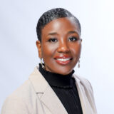 A person with short dark hair wearing a black turtleneck and cream-colored blazer poses for a photo in front of a white backdrop