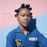 A person with short dark hair wearing a blue jumpsuit poses for a photo against a wall painted pink and blue