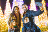 a family posing in winter coats in front of a group of illuminated trees taking a selfie