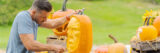 Brown hair man leaning over wearing a grey shirt holding a tool with a sharp edge pointed into an orange pumpkin face that is being carved to look like a human face. A carved out jack-o-laterned with a cut up mouth is in the background of the sculptural looking carved face of the main pumpkin.