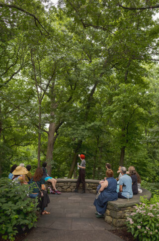 A person in black and white clothing with vivid fuchsia hair gestures to a green-leaved tree while speaking to a sitting tour group