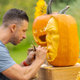 Brown hair man leaning over wearing a grey shirt holding a tool with a sharp edge pointed into an orange pumpkin face that is being carved to look like a human face. A carved out jack-o-laterned with a cut up mouth is in the background of the sculptural looking carved face of the main pumpkin.