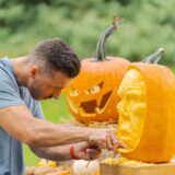 Brown hair man leaning over wearing a grey shirt holding a tool with a sharp edge pointed into an orange pumpkin face that is being carved to look like a human face. A carved out jack-o-laterned with a cut up mouth is in the background of the sculptural looking carved face of the main pumpkin.