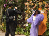 A person in a fluffy purple dress and large hat poses for a photo among yellow flowers