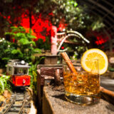 A cocktail in a tumbler sits alongside a model train track in a holiday setting