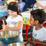 Two children in summer clothes work together to chop and stir vegetables in a bowl