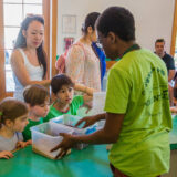 A teenager in a green shirt teaches three children about plant parts in a classroom setting