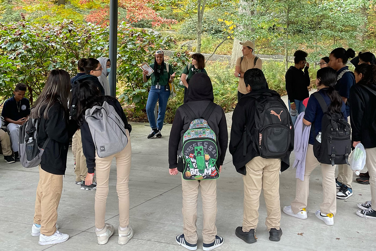 A group of students wearing backpacks listen to a tour guide in a green garden space