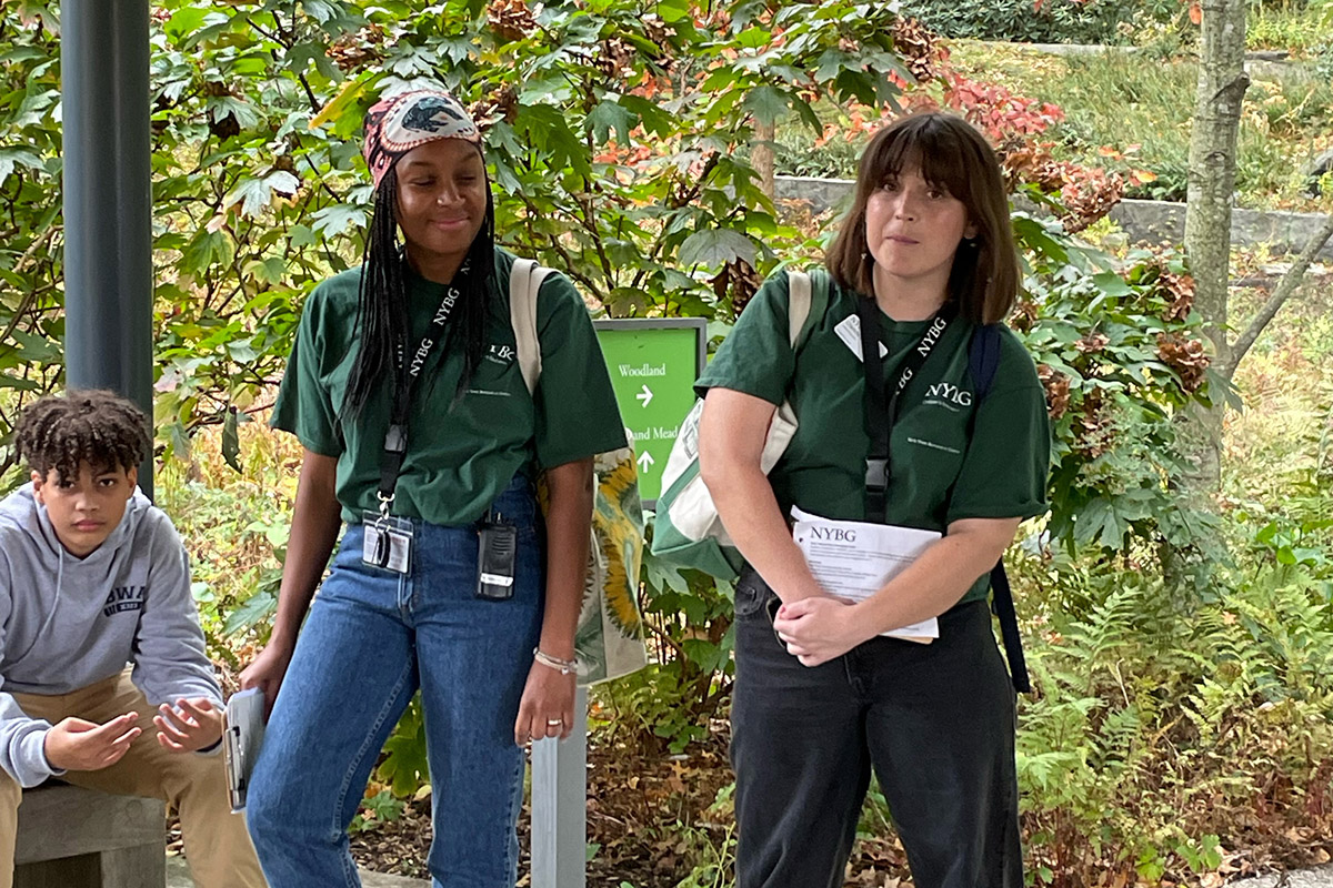 Three students in green shirts pose for a photo in front of a lush garden setting