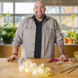 a person stands behind a wooden table in a black shirt and grey long sleeved button down; on the table are yellow orchids and some tools