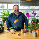 Marc Hachadourian, Orchid Curator, stands behind a wooden table in a blue collared shirt with an orange orchid in front of him and a purple orchid next to him on the table