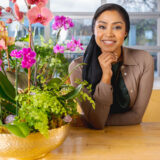 a person in a brown long sleeved shirt and black headwrap leaning on a wooden table with a hand underneath their chin; a pink orchid is positioned to the left in a gold pot