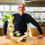 A person in a black shirt and glasses poses for a photo with a brightly lit arrangement of yellow orchid flowers