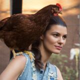 A person in a denim vest poses for a photo with a brown chicken on their shoulder