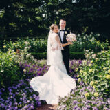 a person is dressed in a white wedding gown holding a bouquet of flowers next to a person in a black tuxedo smiling at the camera surrounded by purple and green flowers