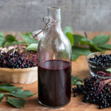 a bottle of dark purple elderberry syrup in a glass bottle, with elderberries around the bottle on a wooden table
