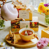 a white teapot pouring tea into a yellow mug; boxes of Tea Forte Paradis tea are in the background