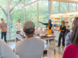 A group of people stands around a table in a brightly lit laboratory, listening to a tour guide