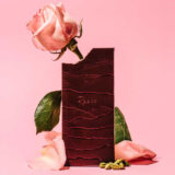 A chocolate bar with a bite taken out of it stands vertically, surrounded by pink rose petals and green nuts