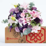 A pink and purple arrangement of roses sitting on a brown and red box labeled 'BloomsyBox'