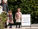 a small child smiles to the camera in a floral jacket and skirt and stands next to a sign