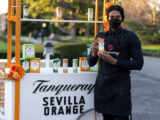 a person in a black button down and black mask holds up a can of Tanqueray mixed drink in front of a stand with "Tanqueray Sevilla Orange" on the front