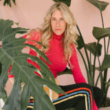 a woman with long blonde hair in a red long sleeved shirt and black pants with colorful stripes on the side sits among green plants