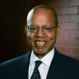 A headshot of a man with glasses in a suit and tie.