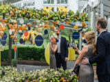 a couple takes a picture of a mirrored display with green and orange florals on top of the display and DeBeers logos on the facade