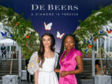 a person in a white dress stands next to another person in a hot pink dress in front of a DeBeers floral display