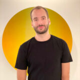 A headshot of a man in a black shirt in front of a background with a yellow circle.