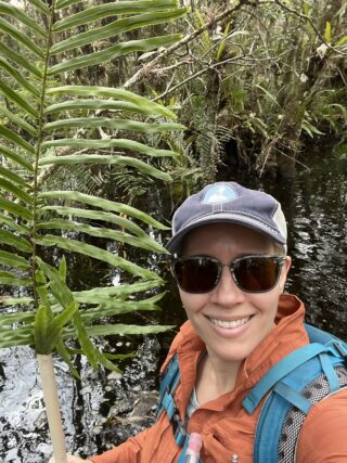 A woman with a blue hat and sunglasses holds up a fern frond.
