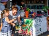 a person holds a blender full of fruits and vegetables as a child pours in milk into the blender