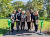 A group of individuals standing behind a stanchion, right after one of them cut the ribbon with a pair of large scissors. The individuals are holding the ribbon and in front of grass with large trees behind them, with upturned lips.