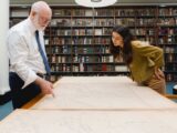 A person wearing a white button down shirt and blue tie, points to a large map on a table. There is a wall with many books on display in the background. Another person on the other side of the table looks over with their hands placed behind them looking down at the map.