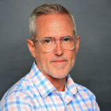 A headshot of a man with glasses and a light blue and pink plaid shirt on.