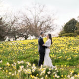 Two people in wedding clothes embrace among a field of white and yellow flowers