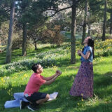 A person in a pink shirt kneels in a field of white and yellow flowers to propose to their partner on a spring afternoon