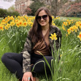 A person in a black jacket and sunglasses sits among a profusion of orange and yellow flowers while posing for a photo