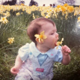 A toddler sits among a field of yellow flowers, trying valiantly to eat one of them