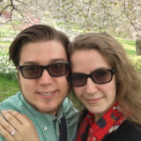 Two people in sunglasses pose for a photo beneath a sunny tree full of pink flowers