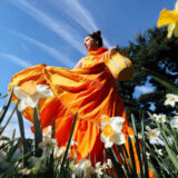 A person in a bright orange dress moves while snapping a photo among white and yellow flowers