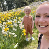 A parent and child smile for the camera while standing in a sunny field of white and yellow flowers