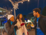 Three people in winter clothes enjoy drinks during an evening event