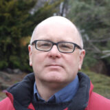 A photo of a person with black glasses and a red coat posing for a headshot