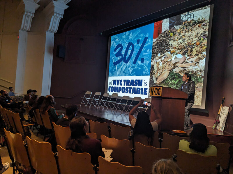 A person stands behind a podium while gesturing at a projector screen, presenting to a room of people